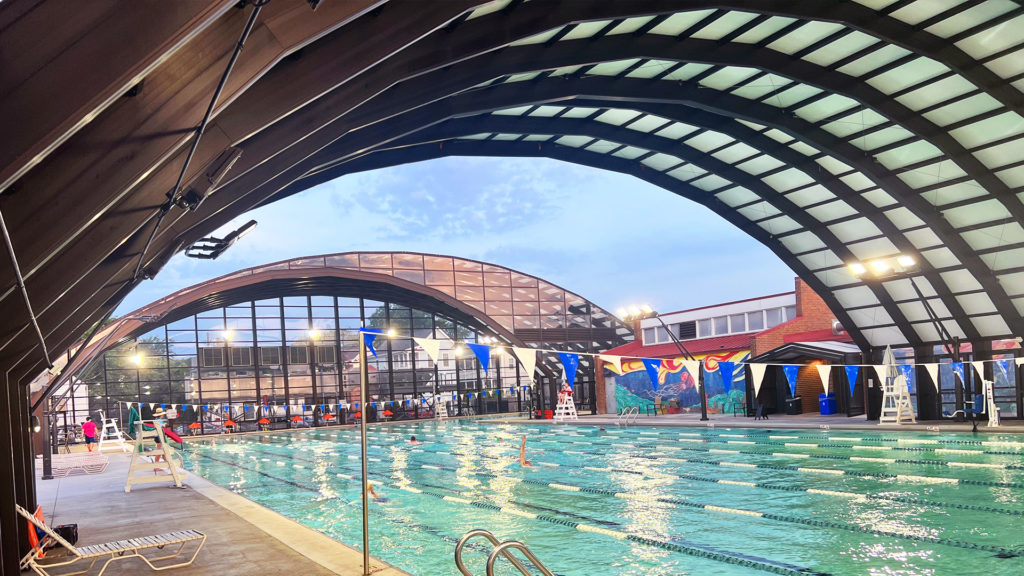 DynaDome | A retractable enclosure swimming pool under a building at dusk.